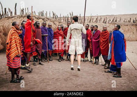 JUN 24, 2011 Serengeti, Tanzania - Group of African Masai or Maasai tribe man in red cloth standing by clay hut in village and European tourist jumpin Stock Photo