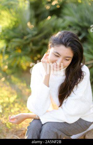 Japanese girl in white sweater sitting on grass in park. Stock Photo
