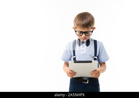 Little boy in black glasses with transparent glasses, blue shirt, pull-ups, blue pants looks into the tablet Stock Photo