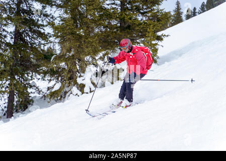 Side image of sports man in red jacket and with backpack skiing in winter resort from snowy slope with trees Stock Photo