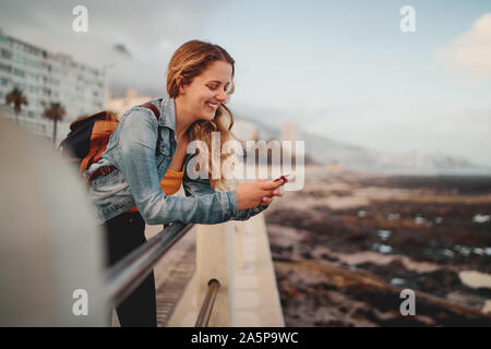A smiling blonde young female traveler with her backpack leaning on railing at city street using her mobile phone Stock Photo