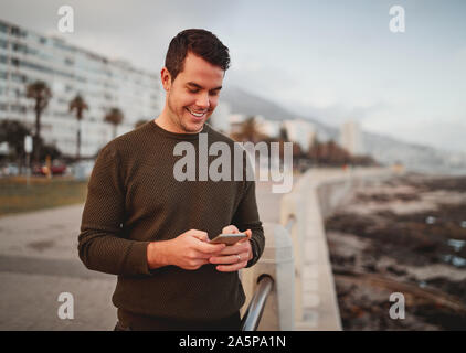 Portrait of a happy attractive young man standing on city street texting massages on his smart phone Stock Photo