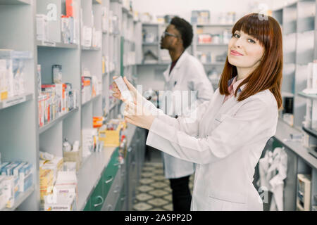 Two Pharmacists Working. Caucasian Woman and African Man Wearing Special Medical Uniform. Woman Showing package with Pills. Man looking for medicines Stock Photo