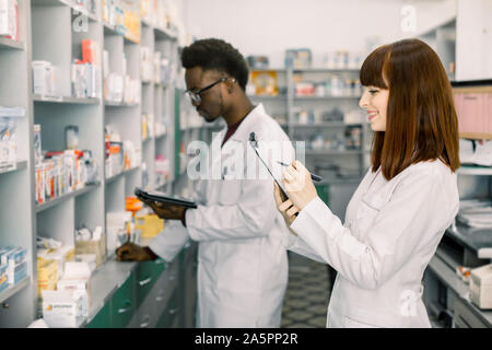 Confident Male And Female Pharmacists In Pharmacy. African American male pharmacist working on digital tablet and Caucasian woman making notes on