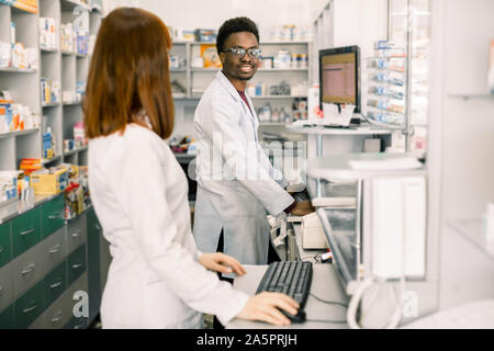 Female and male pharmacists in modern pharmacy, looking at each other, while working on computers Stock Photo
