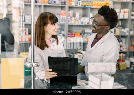Young woman Pharmacist Writing On Clipboard While her Colleague African man watching at her and talking Stock Photo