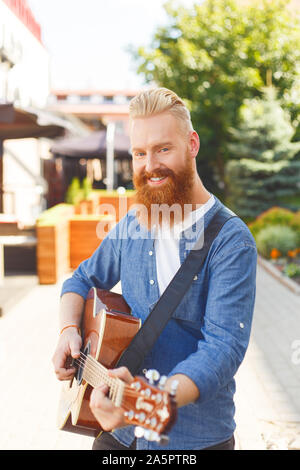 Young bearded man wearing blue Denim jacket, playing guitar outdoors Stock Photo