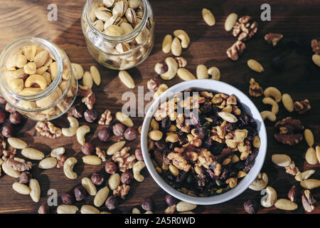 top view of a white bowl with varied organic dry fruits and glass jars with mixed nuts on rustic wooden table, healthy food and snack concept