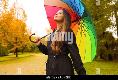 Portrait of a happy woman wearing raincoat under an umbrella breathing in city park on rainy day. Stock Photo
