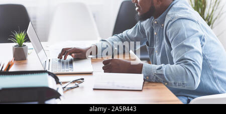Black trainee taking notes while using laptop in office Stock Photo