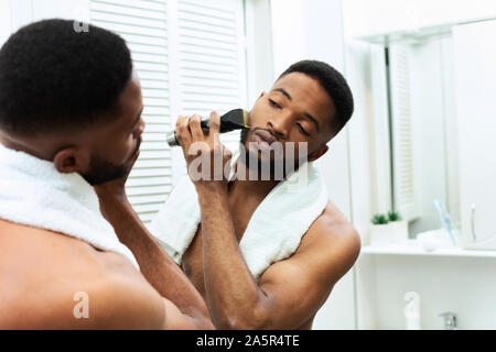 Young black man straightening his beard with trimmer at bathroom Stock Photo