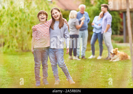 Laughing siblings on the lawn in the garden with family in the background Stock Photo