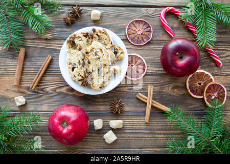Traditional Christmas stollen cake. German festive dessert on brown wooden table background. Red apples, cinnamon sticks, dried orange slices decorati Stock Photo