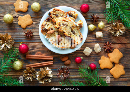 Christmas traditional cake stollen on brown rustic table. Gingerbread cookies, red christmas balls and cinnamon spices decorations. Golden cones with Stock Photo