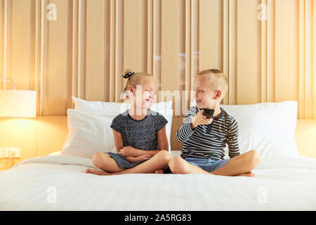 Smiling little brother and sister looking at each other while sitting on a bed watching television together Stock Photo