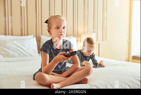 Little girl sitting on a bed looking bored while watching television with her brother lying behind her using a digital tablet Stock Photo