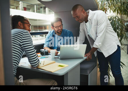 Diverse group of smiling creative professionals working together on a laptop in an office meeting pod Stock Photo