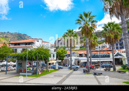 Ribeira Brava, Madeira, Portugal - Sep 9, 2019: Promenade of the Madeiran city photographed from the Atlantic ocean coast. People on the street. Outdoor restaurants, bars, and shops. Palm trees. Stock Photo
