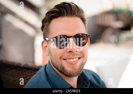 Portrait of young handsome man wearing sunglasses and trendy hairstyle. Businessman smiling and looking into camera outdoors in a cafe. Stock Photo