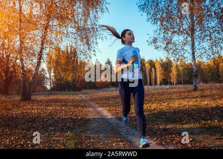 Runner training in autumn park. Woman running with water bottle and keeping fit at sunset. Active lifestyle. Workout
