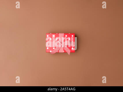 Cute red gift box with white dots and striped ribbon and bow on a brown background. Above view of Christmas gift with a bow. Minimal birthday gift. Stock Photo