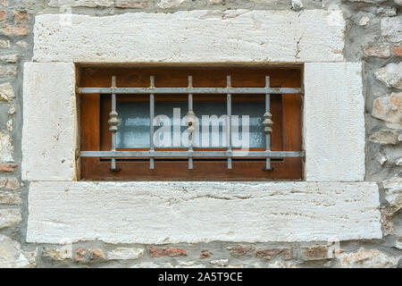 Small window with a brown wooden frame on the stone wall of the house, closed by a metal grill. From a series of windows of the world. Stock Photo