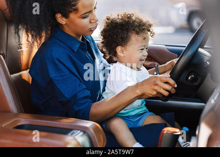 Shot of an adorable little boy sitting on his mother's lap and holding steering wheel. Mother playing with her son in car. Stock Photo