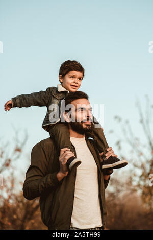 Little boy sitting on father's shoulders on blue sky background.