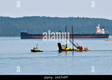 A tug boat towing a barge with an industrial crane across the surface of the water in the Stewart Channel dwarfed by the ocean going ship Stock Photo