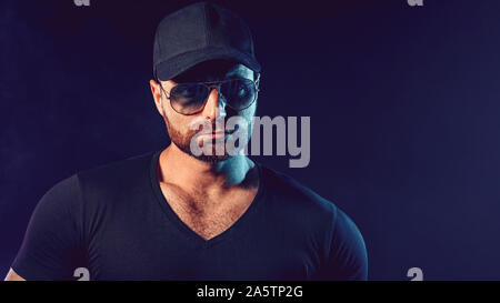 Portrait of brutal bearded man in sunglassesand baseball cap at dark photo studio. Stock Photo