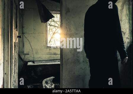 An eerie spooky hooded figure standing by a window in a ruined, abandoned house. With light coming from the winter sun. With a dark moody edit. Stock Photo