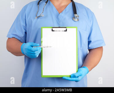 doctor in blue uniform and latex gloves holds a green holder for sheets of paper and with a metal pen shows on a sheet, white background Stock Photo