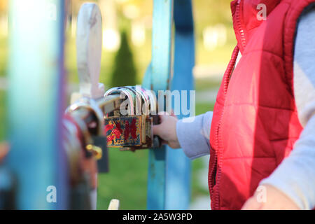 white blond kid in a red waistcoat considering padlocks on the railing of the bridge over the river. Walk in the autumn park Stock Photo