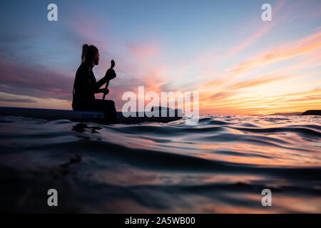 A woman sits and paddles on an undulating ocean during a vibrant sunset afterglow. Stock Photo