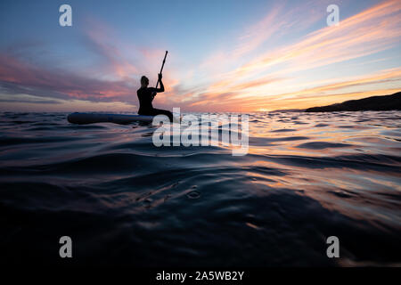 A woman sits and paddles on an undulating ocean during a vibrant sunset afterglow. Stock Photo