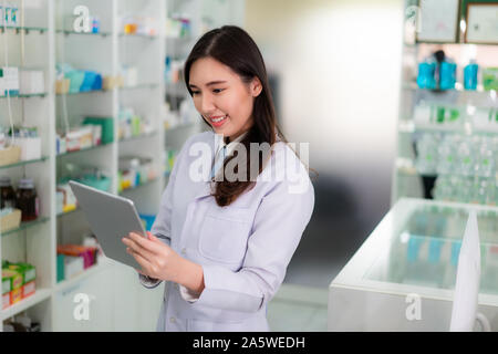 Confident Asian young female pharmacist with smile holding digital tablet and searching that product database in the pharmacy drugstore. Medicine, pha Stock Photo
