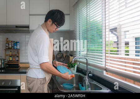 Happy Asian young man in white shirt standing and washing dishes in sink on the kitchen. Kitchen ornaments and kitchenware supplies in background. Man Stock Photo