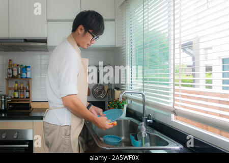 Happy Asian young man in white shirt standing and washing dishes in sink on the kitchen. Kitchen ornaments and kitchenware supplies in background. Man Stock Photo