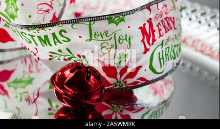 Red Jingle Bell and Decorative Merry Christmas Ribbon with the Words Love & Joy in Red and Green on Silver Reflective Surface Stock Photo