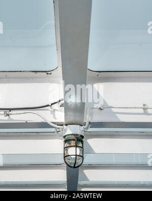 Detail of marine light fixture from underneath, attached to painted white metal beams aboard a ship with view of pale blue sky through ceiling panels. Stock Photo