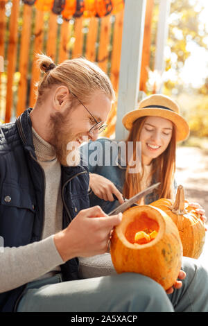 Halloween Preparaton Concept. Young couple outdoors making jack-o'-lantern carving pumpkins smiling joyful side view close-up Stock Photo