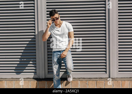 Outdoors Leisure. Stylish guy standing on the street leaning on wall taking off eyeglasses posing to camera cool Stock Photo
