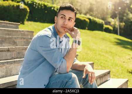 Outdoors Leisure. Stylish guy wearing watch sitting on stairs in park touching face looking aside pensive close-up Stock Photo