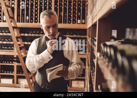 Sommelier Concept. Senior man standing with notebook checking wine collection pensive Stock Photo