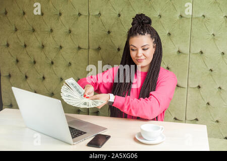 Portrait of successful attractive young woman with black dreadlocks in pink blouse is sitting, holding many dollars, giving you and looking at webcame Stock Photo