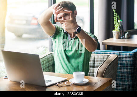 Young serious businessman in green t-shirt sitting with laptop and looking at camera with focus crop composition gesture. business and freelancing con Stock Photo