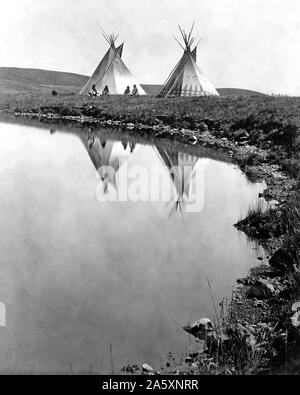 Edward S. Curits Native American Indians -  Two tepees reflected in water of pond, with four Piegan Indians seated ca. 1910 Stock Photo