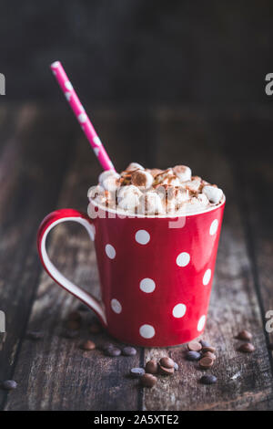 A red and white spotted mug filled with hot chocolate. The drink has whipped cream and mini marshmallows on top, and is on an old wooden table. Stock Photo