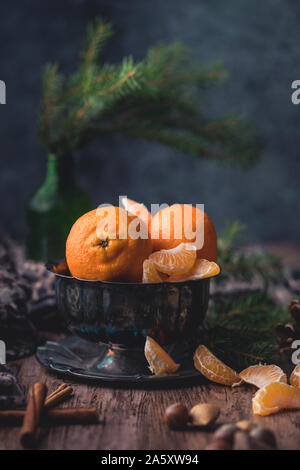 Still life citrus fruits in a metal bowl. Next to bowl are clementine slices, cinnamon sticks and some nuts. There is a green spruce branch in an old Stock Photo