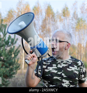 Portrait of a young man screaming with a megaphone in the forest. Lost People Search Concept. Outdoors Stock Photo